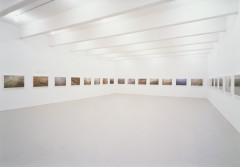 Different color-toned landscape photographs of the Thames River hang in an evenly space, eye-level line along the walls of a white gallery room. The photographs are various close-up detail shots of the water surface.