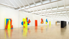 A very large gallery with wood floors, white walls and vertical skylights. Spanning much of the length of the visible wall are 6 large angular paintings, each comprised of shapes painting in blue, yellow and red. in the distance on the right are a series of six identical black slabs or boxes.