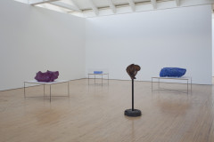 Four evenly spaced objects are on stands in a white gallery room. Three objects in the foreground are organic forms in varying colors of magenta, brown, and blue, and the fourth object in the background is a geometric stack of small, blue prisms under a vitrine.