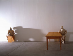Two sculpted figures sit against a white wall. The left figure sits on a short wooden bench with back facing the right figure. The right figure sits parallel to a long wooden table and looks at the left figure's back. A light coming from the left shines onto the left figure and casts a long shadow onto the wall.