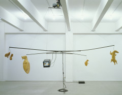 A rudimentary motorized carousel made of exposed poles and cord hangs from the ceiling of a white room. On the carousel hangs five yellow animal forms and a mini color television.