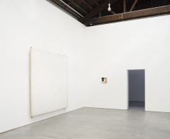 A large white painting and a small work of yellow, black, and white respectively hang on the left and right walls in a white room's corner. Positioned to their right is an open doorway.