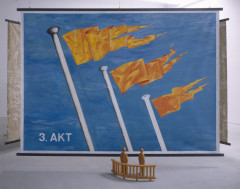 A large painting of orange flags on a blue background hangs from the ceiling. In front of it sits a small wooden sculpture depicting two people standing in front of a fence.