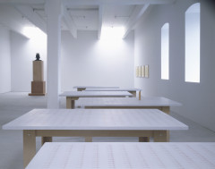 Six wooden tables with white tabletops with a pattern of pink crosses on them are arranged in a slight zigzag shape in a white room with arched windows. A sculpture on a plinth and four framed drawings are in the background.