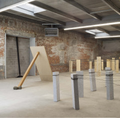 Three sculptures sit in an industrial space with a metal ceiling. From left to right: a wooden plank with rocks at the bottom props up a strip of lighter wood; separate marble planks are held upright by a network of wires; wooden planks are held upright by a network of metal strips.