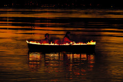 A burning boat on a body of water, reflecting black and orange, with two people sitting in it, one rowing.
