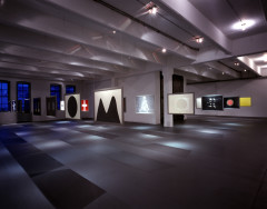 Several images in silver frames—including a black circle on a white background, a white cross on a red background, and more—hang from the ceiling in a dark room, their shadows forming a rectangular pattern on the floor.