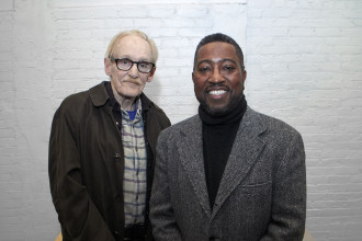 An man wearing glasses and a brown coat over a brown plaid shirt stands next to a smiling man wearing a black turtleneck and grey tweed coat in front of a white brick wall.