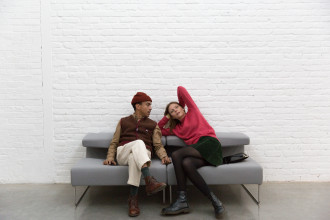 Two visitors sit on a grey plush bench, the one on the left wearing brown and white crosses their legs and looks at the one on the right wearing pink and green and reclining on one elbow with the other in the air and behind their head.