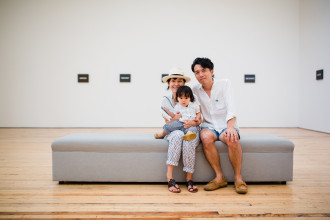 Two adults and a small child sit closely together in a gallery on a grey bench in front of a series of On Kawara's 