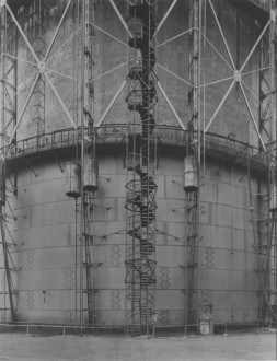 Close-up black-and-white photograph of a gas tank, showing geometric external ladders, staircases, and metal beams.