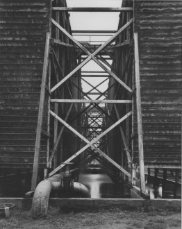 Black-and-white photograph of two building structures on stilts above a pool of liquid flanking an open central passageway filled with sets of diagonal crossbeams extending into the background.