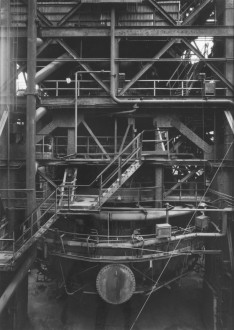 Black-and-white photograph of a tiered metal structure with tubes, railings, and a staircase surrounding a blast furnace.