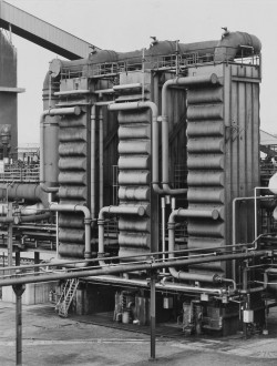 Black-and-white photograph of three vertical cooling towers with a vertical stack of half-cylindrical forms attached to the front and intersecting tubes connecting each tower.