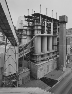 Black-and-white photograph of a windowless building below eight cylindrical towers, topped with scaffolding and tall ventilation tubes within a larger industrial complex.