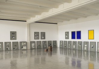 Framed photographs lean against a wall and in a corner. Seven frames with various photographs or blue or yellow paintings are installed above in addition to two stacks of copper and iron blocks.