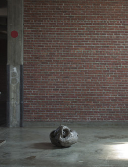 A gray, round sculpture sits on a concrete floor in front of brick wall.
