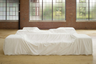 A large-scale, couch-like object is covered with a soft white material in a room with wooden floors and large square windows.