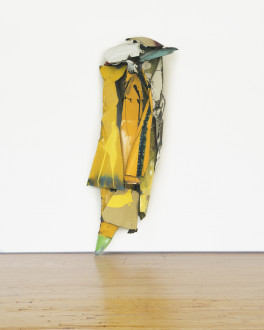 A vertical sculpture composed of curved yellow and orange metal is placed against a white wall.