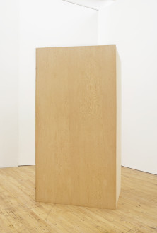 A wooden rectangular box is vertically placed on a wooden floor in a white room.