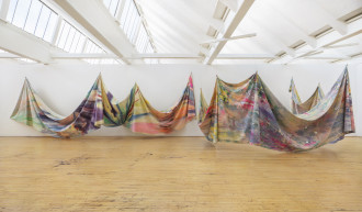Two massive swathes of multicolored canvas are tied at various points and strung up from the ceiling of an airy, bright, white room. The canvases are painted and splattered with an array of vibrant, pastel, and metallic colors. The artwork floats above the floor. The left swathe of canvas is pinned to the wall at the bottom left, center, and right edges. The right swathe of canvas forms a semicircle with the arc facing away from the wall.