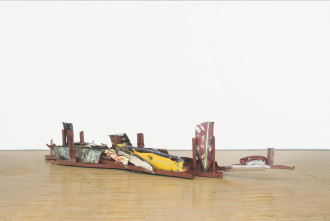 A long, low-lying sculpture made of red, white, yellow, and multicolored metal parts rests on a wooden floor.