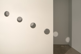 A lit up plaster divider has four identical, evely spaced circles cut out of it. The circles descend from left to right. Circular light silhouettes are cast onto the background wall.