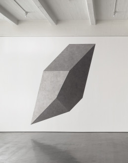 White wall with a central gray shape composed of several geometric shapes in a slightly different shade above a concrete floor. 
