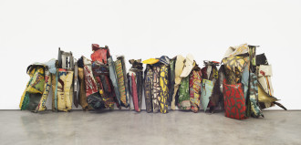 Many crumpled-and-folded metallic objects that have been painted in various colors and patterns are lined up, placed on a cement floor, and rest against a white wall.