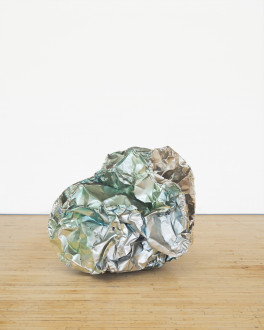 A crumpled ball-like sculpture made of reflective silvery green metal rests on a wooden floor.