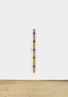 A tall, narrow orange pole with small white and gray rectangles dotted on its surface hangs on a white wall.