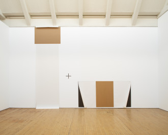 A vertically oriented, rectangular, white-and-brown work on paper is placed near the ceiling and stretches along a white wall down to a wooden floor. Next to it is a horizontally oriented, rectangular, white, brown, and black work on paper that is placed on the wall just above the floor. A plus sign is placed on the wall between the two works on paper.