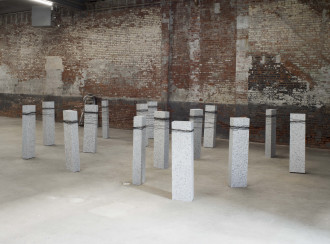 Fifteen upright, rectangular blocks of speckled granite are bound together with steel wire. The sculpture sits on a concrete floor in front of a brick wall.