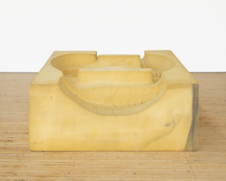 A foam square with curved forms carved into its top rests on a wooden floor in front of a white background.