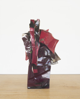 A columnar sculpture made of red metal parts that twist together rests on a wooden floor.