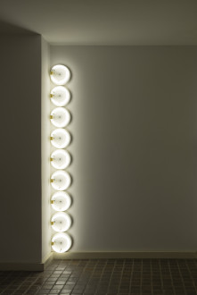 Nine circular, white, fluorescent lightbulbs are placed vertically in the corner of a room.