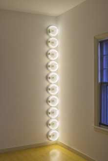 Ten circular, white, fluorescent lightbulbs are placed vertically in the corner of a room.