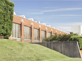Exterior of an industrial brick building with a grassy hill in the foreground and the edge of a row of hedges.