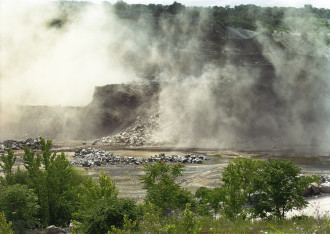 Smoke or fog swirls around and obscures the view of a cliff's edge that is positioned above a river. Lush trees are present in the foreground of the photograph as well as in the distance beyond the cliff's edge.