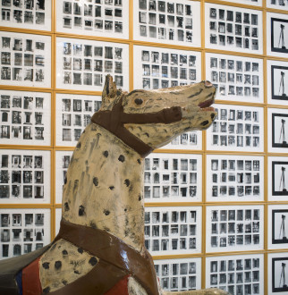 A painted and wooden toy horse in front of a locked grid of paper works, each an individual and unique grid of black and white photographs.
