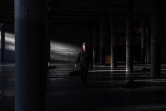 A person in black clothes and a grey beanie walks forwards in a dark industrial basement and is illuminated by natural light.