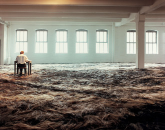 A room with the floor covered in a thick layer of brown animal fur, with a figure seated at a small desk on the left hand side, facing a white wall of windows.
