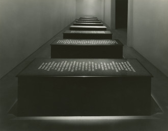Ten black, rectangular marble platforms with all caps text carved into the top face of the platform sit in a single line in a narrow room. Each platform has a light shining on its face illuminating the text.