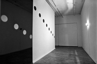 A black and white image of a thin plaster divider sits in the middle of a room. The divider runs ceiling to floor and has eight identical, evenly spaced circles punched out of it. The circles descend from left to right at a slight angle. A light fixture attached to the right wall shines through the circle cutouts and casts light silhouettes onto the left wall. A simliar light fixture is attached to the left wall but is not on.