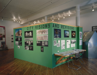 A green rectangular structure in the center of a room, with a desk and chair built into one side. On the walls are hung a variety of photos, diagrams, charts, texts, and other images, with the words 