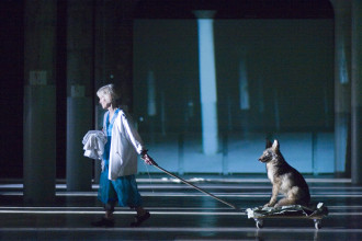 A woman carrying white fabric in one hand drags a dolly with a coyote seated on it through an industrial space lit by blue light and projections.