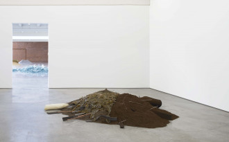 A large, mixed mound of dirt, thick felt strips, shiny gunk, and thin protruding cooper and steel pipes and wire sit on the concrete floor in a brightly lit white room. In the background through a doorway is three other mounds of blue glass, sand, and rocks.