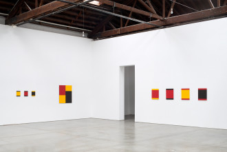 Different square-shaped, color-blocked paintings of red, marigold, and black hang on adjacent white walls.