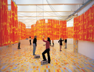 A roomful of red, orange, and yellow curtails hang halfway down from a wavy ceiling track. Figures in the room interact with the curtains and some stand on sporadically placed cinderblocks that are under a microphone hanging from a ceiling cord.