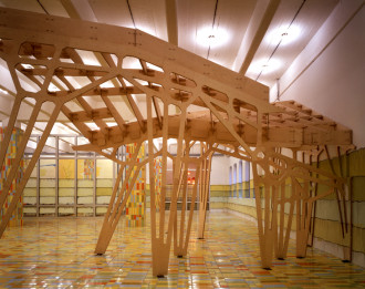 A large wooden prototype model on tall legs fills a large room and almost reachs the ceiling.